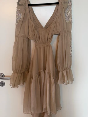Beige kortklänning med pärldetaljer - Romantisk beige kortklänning i luftig chiffong. Klänningen har långa, vida ärmar med vackra broderier och pärlor på axlar och ärmar. Snygg cut-out i ryggen. Perfekt för dig som gillar boho vibes och unika detaljer.