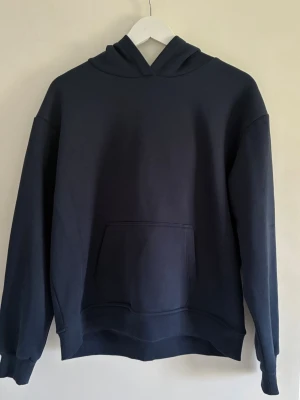 Mörkblå hoodie  - En stilren mörkblå hoodie med klassisk huva och stor magficka framtill. Hoodien har ribbade muddar vid ärmslut och nederkant, samt en avslappnad passform. Perfekt för en chill och casual look. Står M men passar S