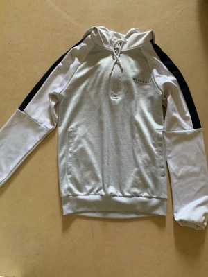Grå hoodie från McKenzie med dragkedja - Snygg grå hoodie från McKenzie med vita och svarta detaljer på ärmarna. Tröjan har huva med dragsko, halv dragkedja framtill och ribbade muddar. Perfekt för en sportig och avslappnad stil.
