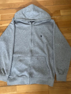 Grå zip hoodie från Polo Ralph Lauren - Snygg gråmelerad hoodie med dragkedja från Polo Ralph Lauren. Klassisk huva, två stora fickor fram och ikonisk liten logga broderad på bröstet. Perfekt för chill dagar och enkel att matcha med allt. Mjuk bomullsblandning och ribbade muddar.