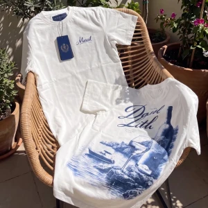 Vit t-shirt med blått tryck - Säljer en vit t-shirt med blått tryck framtill och texten 'Dani Lith'. T-shirten har klassisk passform, rund hals och korta ärmar. Materialet känns mjukt och skönt, perfekt för sommardagar. Trycket visar en vinflaska, glas och havsmotiv för en chill vibe.
