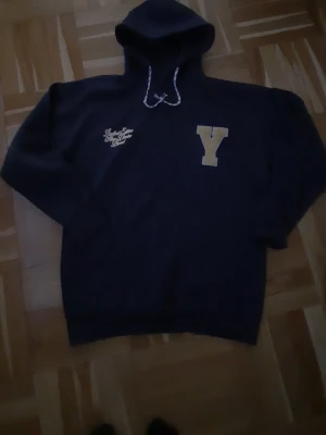 Mörkblå hoodie med gul brodyr - Säljer en mörkblå hoodie med huva och vita dragsnören. På bröstet finns ett stort gult Y och broderad text i gult som säger 'Limited Edition New Denim Brand'. Perfekt för dig som gillar college-stil och vill ha något bekvämt och snyggt. S i men