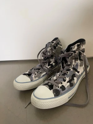 Converse  - Snygga high-top sneakers med svart, grå och vit camo-mönster. Skorna har klassisk gummisula, vita tåkappor och grå skosnören.