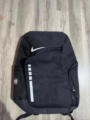 Svart Nike Elite ryggsäck - Snygg svart Nike Elite ryggsäck med vit logga och reflekterande detaljer. Ryggsäcken har flera fack, justerbara axelband med ELITE-tryck och robust dragkedja. Perfekt för skola, träning eller vardagsbruk. Tillverkad i slitstarkt syntetmaterial.
