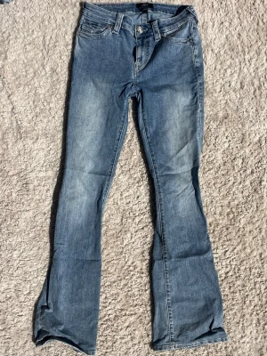 Blå bootcut jeans från True Religion - Säljer ett par ljusblå bootcut jeans från True Religion med klassiska kontrastsömmar och detaljerade bakfickor. Jeansen har normal midja och är tillverkade i denim med lätt tvättad look. 