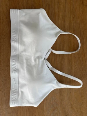 Vit sport-bh från Nike - Snygg vit sport-bh, Nike Indy, med bred resår där Nike-loggan är präglad runt om. Bh:n har smala axelband och är tillverkad i ett mjukt, stretchigt material som ger bra stöd och komfort. Perfekt för träning eller en aktiv vardag.