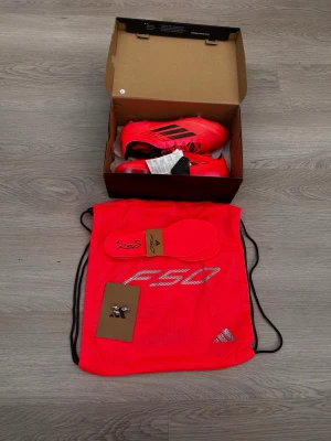 Adidas F50 röda sportskor - Riktigt snygga Adidas F50 sportskor i rött med svarta detaljer och klassiska tre ränder på sidan. Skorna är låga och tillverkade i lätt syntetmaterial. Medföljer extra innersulor och en matchande röd gympåse med F50-tryck.