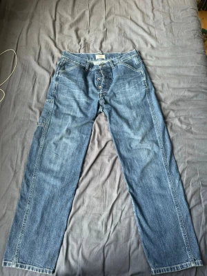 Tela Genova Heritage DAVIDE/3F – IL MARE - Säljer ett par klassiska blå jeans med raka ben och knappgylf. Jeansen har fem fickor och tydliga sömmar längs benen. Materialet är denim i bomull och färgen är en tvättad mellanblå nyans. Perfekta för en avslappnad stil. 