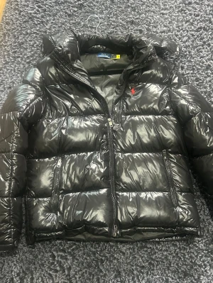 Svart pufferjacka från Polo Ralph Lauren - Säljer en svart, glansig pufferjacka från Polo Ralph Lauren med röd logga på bröstet. Jackan har hög krage, dragkedja framtill och två sidofickor med dragkedja. Perfekt för kalla vinterdagar och riktigt snygg streetstil.