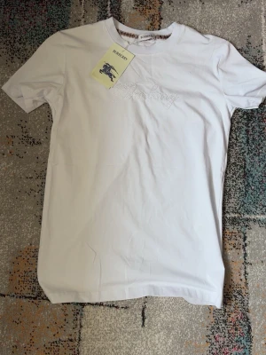Vit Burberry t-shirt med tryck - Snygg vit t-shirt från Burberry med pärlbroderad logga framtill. Klassisk rund halsringning och korta ärmar. Tillverkad i mjuk bomull som känns skön mot huden. Perfekt för dig som gillar stilrena plagg med exklusiva detaljer.
