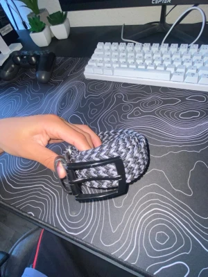 Svart och grått flätat bälte - Säljer ett snyggt flätat bälte i svart och grått. Bältet har ett klassiskt metallspänne och är gjort i ett elastiskt syntetmaterial. Perfekt för att ge din outfit en cool touch.