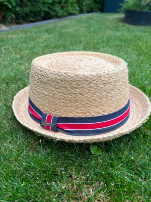 Wigéns stråhatt strl L - Snygg stråhatt i naturfärg med ett dekorativt band i blått och rött. Hatten har en klassisk rak form och är tillverkad av flätat stråmaterial. Perfekt för soliga dagar och ger en cool touch till din outfit.