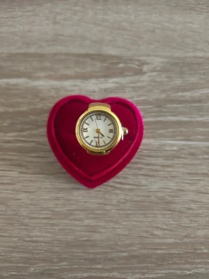 Guldfärgad ring med klocka - Unik guldfärgad ring med inbyggd klocka och vit urtavla med romerska siffror. Ringen har en klassisk rund form och detaljerad urtavla. Perfekt för dig som vill ha något annorlunda och iögonfallande bland dina accessoarer.