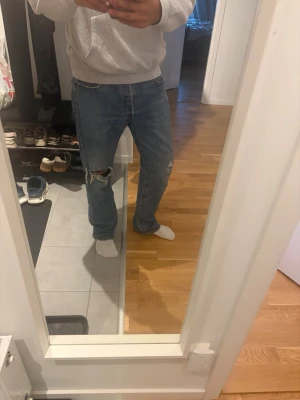 Levi's blå raka jeans 501 - Säljer ett par klassiska blå Levi's jeans med raka ben och slitningar vid knät. Jeansen har fem fickor, gylf med knappstängning och den ikoniska läderlappen bak. Perfekta för en avslappnad och trendig look.
