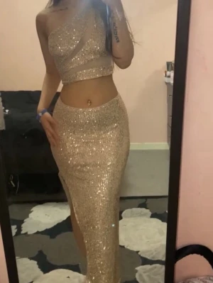 Glittrigt guldigt set med slits - Säljer ett glittrigt tvådelat set i guld med en axelbandslös crop top och en lång kjol med hög slits. Setet är täckt av paljetter och har en figurnära passform som framhäver midjan. Perfekt för dig som vill sticka ut och glänsa.