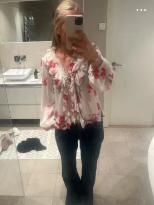 Vit blommig blus med volanger - Söt vit blus med rosa blommor och volangdetaljer längs ringningen och framtill. Blusen har långa ärmar med resår vid handlederna och spetskant vid halsen. Luftigt material som ger en romantisk känsla.