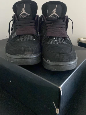 Svarta Air Jordan 4 sneakers - Säljer ett par svarta Air Jordan 4 sneakers med meshdetaljer och Jumpman-logga på plösen och sulan. Skorna har svarta skosnören och en robust sula med tydligt mönster. Materialet är en mix av mocka och textil, vilket ger en klassisk streetwear-look.