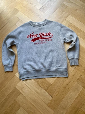 Grå sweatshirt  - Säljer en grå sweatshirt från ZARA med rött New York-tryck och texten 'State of Mind Since Forever' på bröstet. Tröjan har rund hals, långa ärmar och ribbade muddar. Perfekt för en avslappnad och cool stil.