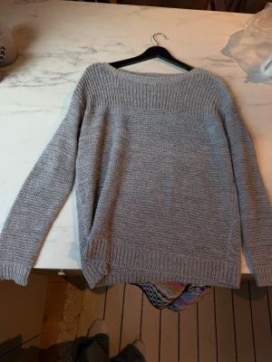 Ljusgrå stickad tröja - En enkel och stilren ljusgrå stickad tröja med rund halsringning och långa ärmar. Tröjan har ribbstickade muddar vid ärmslut och nederkant samt en diskret metallbricka nere vid fållen. Perfekt för lager på lager under kyliga dagar.