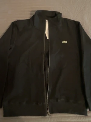 Svart zip hoodie från Lacoste - Säljer en svart hoodie från Lacoste med dragkedja framtill och klassisk ståkrage. Tröjan har det ikoniska gröna krokodilmärket på bröstet och är tillverkad i mjukt bomullsmaterial. Långärmad och enkel design som passar till det mesta.