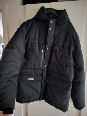 Svart dunjacka från Geographical Norway - Snygg svart dunjacka från Geographical Norway med huva, flera stora fickor framtill och dragkedja. Jackan har patchdetaljer och tryckknappar, samt en norsk flagga på ärmen. Perfekt för kalla vinterdagar och riktigt varm.