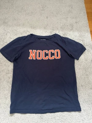 Mörkblå NOCCO t-shirt - Snygg mörkblå t-shirt med NOCCO-tryck i vitt och orange på bröstet. Klassisk passform och rund halsringning. Perfekt för dig som gillar enkel och sportig stil.