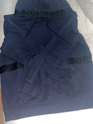 Mörkblå hoodie från HUGO - Snygg mörkblå hoodie från HUGO med svart logotypband på huvan och ärmarna. Klassisk känguruficka framtill och justerbar dragsko i huvan. Tillverkad i mjukt bomullsmaterial som känns skönt mot huden. Perfekt för en avslappnad och trendig look.