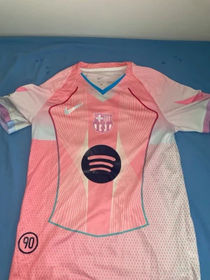 FC Barcelona rosa matchtröja Nike - Rosa och vit FC Barcelona matchtröja med kort ärm, V-ringning med blå kant och tryckta detaljer. Framsidan har klubbmärke, Nike-logga och Spotify-logga. Baksidan har texten Lamine Yamal och nummer 10. Tillverkad i lätt och ventilerande material, perfekt för fotboll.