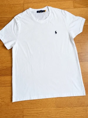 Vit t-shirt från Polo Ralph Lauren - Klassisk vit t-shirt från Polo Ralph Lauren med rund hals och korta ärmar. T-shirten har den ikoniska broderade loggan i marinblått på bröstet. Tillverkad i mjuk bomull för en skön känsla och enkel stil.