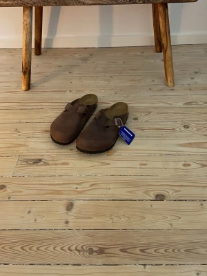 Bruna tofflor i mocka med rem - Säljer ett par bruna tofflor i mocka med justerbar rem över foten och svart räfflad sula. Tofflorna har en klassisk slip-in design och innersulan ser ut att vara mjuk och bekväm. Perfekta för chill dagar hemma eller när du vill ha något enkelt att slänga på dig. Tyvärr ingen skolåda. Aldrig använda då de tydligt är för stora. 