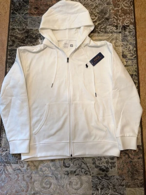 Vit zip hoodie från Polo Ralph Lauren - Snygg vit hoodie med dragkedja från Polo Ralph Lauren. Klassisk design med huva och dragsnören, två fickor fram och den ikoniska lilla loggan broderad på bröstet. Tillverkad i mjukt material som känns skönt mot huden. Perfekt för en clean och stilren look.