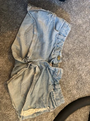 Ljusblå väldigt lågmidjade jeansshorts från Tezenis - Ett par ljusblåa jeansshorts från TEZENIS. Ganska korta och lågmidjade med vit spetts längst sidan och baksidan. Även lite hål på vissa ställen. Har klippt bort storleks lappen men det är i strlk (xs-s). Kan skicka mått om det önskas. 
