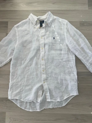 Vit linneskjorta från Polo Ralph Lauren - Klassisk vit skjorta i linne från Polo Ralph Lauren med blå broderad logga på bröstet. Skjortan har knappar framtill, button down-krage och lång ärm. Perfekt för en clean och fräsch look.