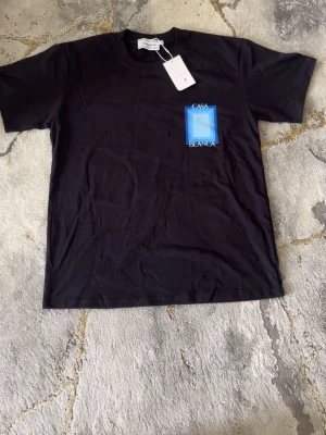 Svart Casa Blanca t-shirt med blått tryck - Snygg svart t-shirt från Casa Blanca med ett stort blått grafiskt tryck och texten 'Casa Blanca' på ryggen samt en mindre version på bröstet. Klassisk passform och rund hals. Perfekt för dig som gillar streetwear och vill sticka ut lite extra.