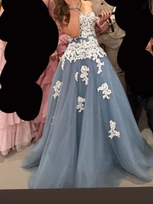Balklänning - Säljer en blå balklänning med vita spetsapplikationer och axelbandslös design. Klänningen har en vid kjol i tyll och vacker v-ringad överdel med blommiga spetsdetaljer. Perfekt för bal eller festliga tillfällen. Ny pris 6000kr använd en gång!! Skriv vid frågor