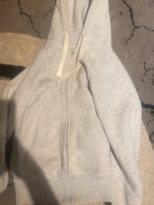 Russedress Hoodie med Zip  - Ljusgrå hoodie med dragkedja och stor huva. Tröjan har fickor framtill och ribbade muddar vid ärmslut och nederkant. På ena ärmen finns ett diskret tryck med texten 'Russedress'. Perfekt för en chill och avslappnad stil.