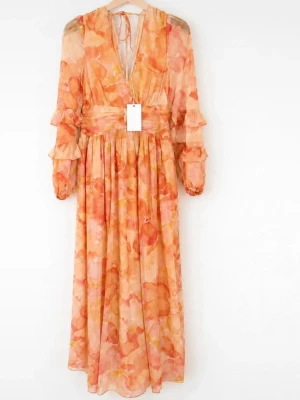 Ny oanvänd Nakd maxiklänning  - Romantisk långklänning med blommigt mönster i orange, rosa och beige toner. Klänningen har v-ringning, markerad midja, volangdetaljer på ärmarna och långa ärmar med resår vid handlederna. Tillverkad i ett lätt, luftigt material som passar perfekt till sommaren.
