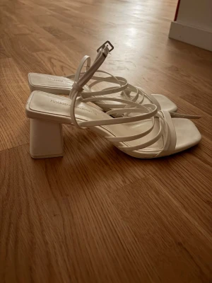 Vita strappy sandaler med klack - Säljer ett par vita sandaler med fyrkantig tå, tunna remmar och bred klack. Sandalerna har justerbart spänne runt ankeln och en stilren design som passar till många outfits. I konstläder.