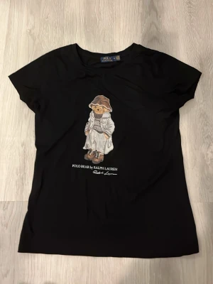 Svart Polo Bear t-shirt från Ralph Lauren - Svart t-shirt från Polo Ralph Lauren med ikoniska Polo Bear-trycket framtill. Klassisk rund hals och korta ärmar. Tillverkad i mjuk bomull för en skön känsla och stilren look.
