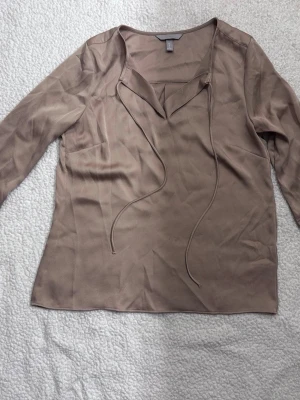 Beige blus med knytband - Stilren beige blus med långa ärmar och v-ringning med dekorativa knytband framtill. Tillverkad i ett mjukt, lätt tyg som ger ett snyggt fall. Perfekt att matcha med jeans eller kjol för en trendig look.