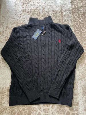  Polo Ralph Lauren - Kabelstickad tröja från Polo Ralph Lauren med halv dragkedja och ribbad krage. Tröjan har det klassiska röda Polo-logot på bröstet och ribbade muddar vid ärmslut och nederkant. Perfekt för dig som gillar stilren och tidlös design. Tröjan är i S men skulle säga att den är mer åt i M