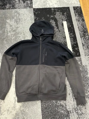 Tvåfärgad hoodie med dragkedja - Säljer en snygg sail racing hoodie i svart och mörkgrått med hel dragkedja framtill. Tröjan har huva med tryckt logga, bröstficka med dragkedja och två sidofickor. Perfekt för chill dagar eller när du vill ha en enkel men stilren look.