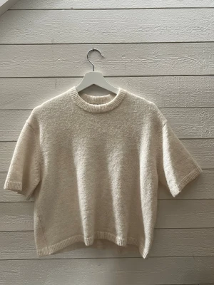 Krämvit stickad tröja Gina Tricot - Krämvit stickad tröja från Gina Tricot med korta ärmar och rund hals. Tröjan har en boxy passform och ribbade detaljer vid hals, ärmslut och nederkant. Tillverkad i ett mjukt, fluffigt material som känns lyxigt och skönt.