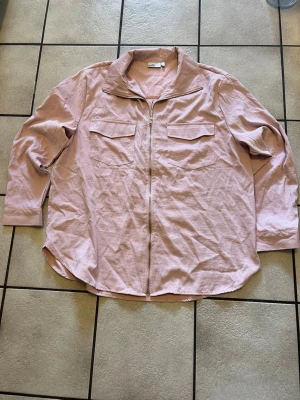Ljusrosa overshirt med dragkedja storlek L - Ljusrosa overshirt i mjukt tyg med dragkedja framtill och två bröstfickor med lock. Avslappnad passform och klassisk krage. Perfekt för lager på lager och enkel att styla till olika outfits.