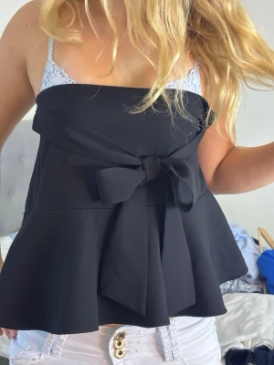 Svart topp med volang och knyt rosett från Zara - Trendig svart topp från Zara med bred volangkant och stor knytrosett i midjan. Toppen har en feminin siluett och är tillverkad i ett mjukt, följsamt material som ger en snygg passform. Jag köpte den secondhand och säljer då den är för liten