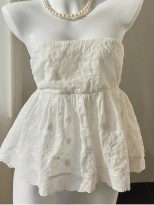 Vit axelbandslös spets tubtopp - Söt vit axelbandslös tubtopp med broderade blommor och spetsdetaljer. Toppen har en lätt utsvängd passform och romantisk känsla, perfekt för sommarens varma dagar. Tillverkad i bomull med fina spetskanter nertill🤍