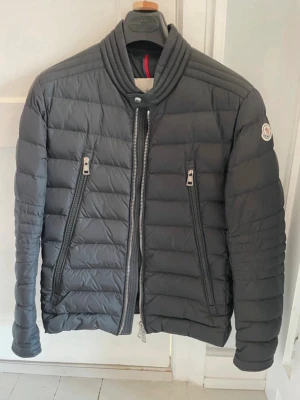 Moncler Down Jacket Size 1 - This is a Moncler down jacket in black color. It features a padded, quilted design with two large diagonal zippered pockets on the front and a central zipper closure. The Moncler logo patch is visible on the left sleeve. The collar is ribbed and stands up for added warmth. The back is plain with horizontal quilting. The jacket appears to be in good condition with no visible stains or tears.
