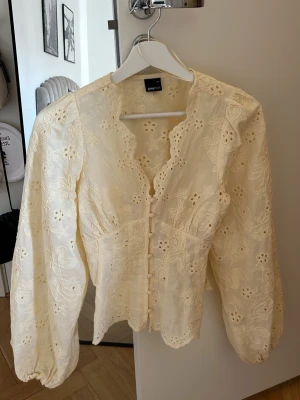 Broderad blus från Gina Tricot - Söt blus från Gina Tricot med broderade blommor, knappar framtill och vågig kant vid halsen. Blusen har puffiga långärmar och är gjord i ett tunt, luftigt material som känns romantiskt och trendigt.