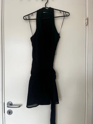 Svart kort klänning från Gina Tricot - Snygg svart kort klänning från Gina Tricot med ärmlös design och smickrande passform. Klänningen har ett band i midjan som kan knytas för extra detalj. Perfekt för dig som gillar stilrena och trendiga plagg.