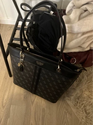 Svart handväska från Guess - Snygg svart handväska från Guess med guldfärgade detaljer och dubbla handtag. Väskan har ett diskret mönster med Guess-loggor och dragkedja upptill. Insidan är ljus med flera fack för smart förvaring. Perfekt för dig som vill ha både stil och funktion.